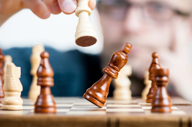 A person's hand holding a white pawn over a chessboard, with a brown bishop lying on its side, indicating a move or capture in a game of chess. The player is out of focus in the background.